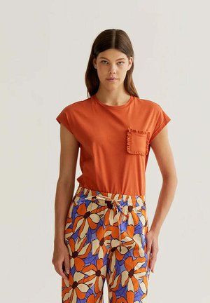 Mujer con una camiseta naranja de manga corta con un bolsillo con volantes y pantalones florales coloridos con pétalos naranjas, crema y púrpuras.