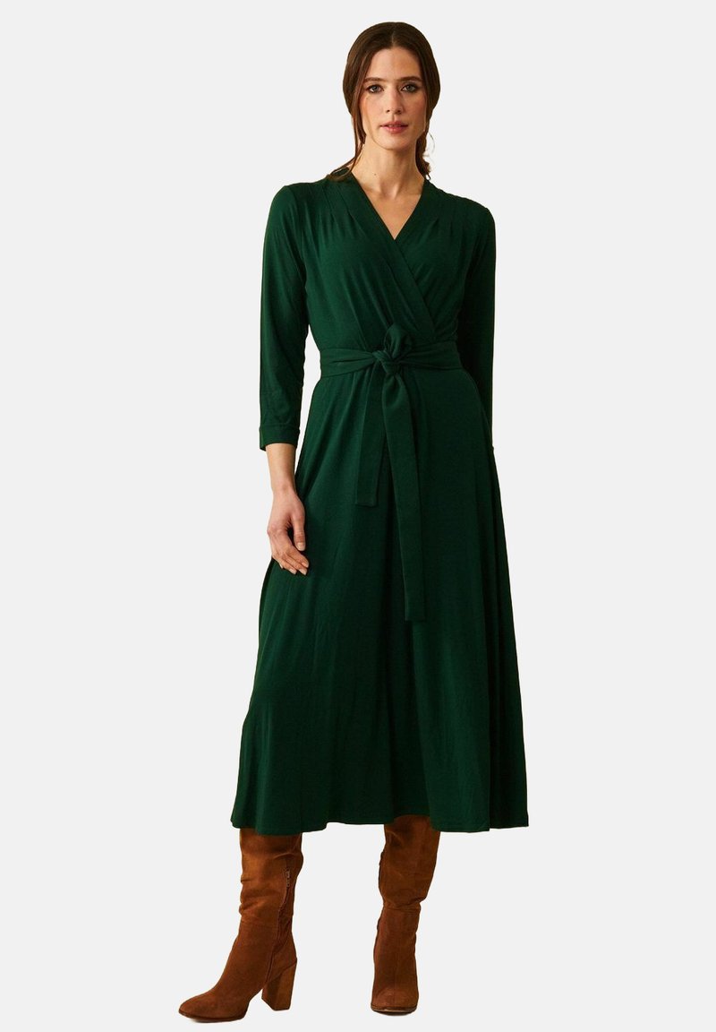 Modell klädd i en grön omslagsklänning med trekvartsärmar, åtsittande vid midjan, tillsammans med bruna knähöga stövlar, mot en enkel bakgrund.