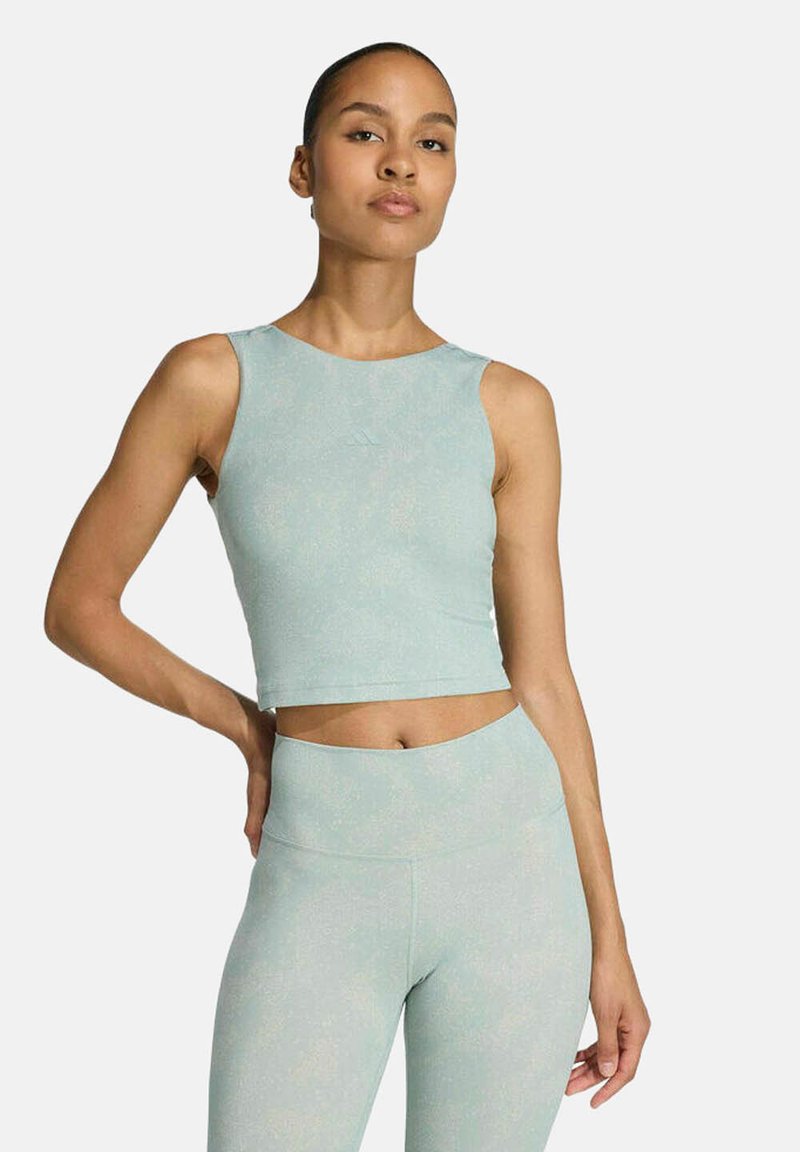 Femme aux cheveux tirés en arrière portant un haut court sans manches bleu clair et un legging taille haute, debout avec une main sur la hanche.