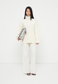 Överdimensionerad krämfärgad ribbad turtlenecktröja, matchad med vita vida byxor. Håller en silverfärgad clutch med ormskinnsmönster och bär svarta sandaler.