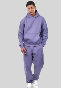 Lila Kapuzenpullover und passende Jogginghose aus weichem Stoff. Verfügt über eine Fronttasche und elastische Bündchen. Getragen mit weißen Sneakers.