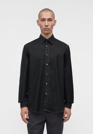 Homme aux cheveux courts portant une chemise noire ajustée à boutons avec des détails de couture blancs et un pantalon sombre, debout sur un fond clair uni.