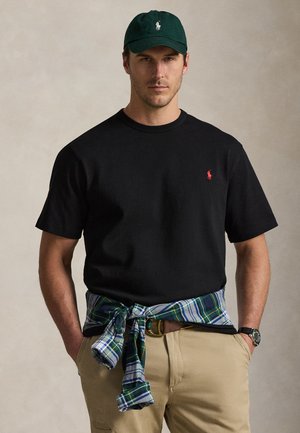 Mand iført sort t-shirt med rødt logo, grøn kasket med hvidt logo, khaki bukser, ternet skjorte bundet om livet og ur på venstre håndled.