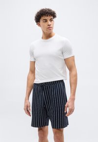 Νεαρός άνδρας με σγουρά μαλλιά, φορώντας απλό λευκό T-shirt και μπλε σκούρο σορτς με λευκές κάθετες ρίγες, κοιτάζει προς τα πλάγια μπροστά σε λευκό φόντο.