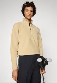 Persoană cu păr scurt și creț poartă un hanorac de fleece bej cu fermoar până la piept și pantaloni negri, ținând bețe de trekking negre cu mânere gri.