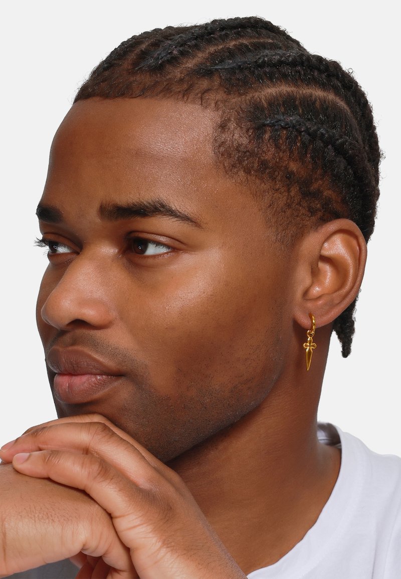 Boucle d'oreille en or avec un charme en forme de dague. Les cheveux de la personne sont coiffés en petites nattes, mettant en valeur les détails de la boucle.