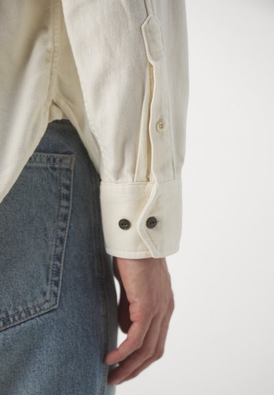 Camisa de botón color crema con un acabado texturizado, que presenta un puño de doble botón, combinada con jeans de denim azul.