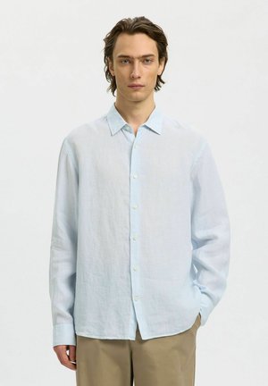 Młody mężczyzna stojący z jedną ręką w kieszeni, ubrany w jasnoniebieską koszulę na guziki i beżowe spodnie na jednolitym tle.