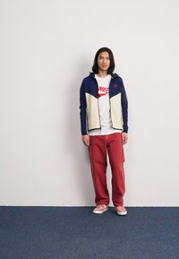 Marineblå og cremefarvet zip-up jakke over en hvid Nike-logo t-shirt, parret med røde bukser og lyse sneakers. Enkel design, afslappet pasform.