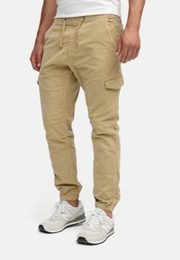 Pantalones cargo beige con cintura elástica, cierre con cordón, bolsillos laterales y puños ajustados. Fabricados en un ligero tejido de algodón.