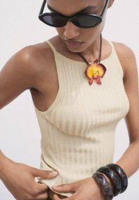 Top in maglia a coste beige con spalline sottili, indossato con occhiali da sole neri e una grande collana con pendente floreale. Bracciali scuri sul polso.