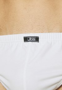Witte katoenen tailleband met een zwart label waarop "jbs." staat. Gevoelige huid is zichtbaar boven de tailleband. Eenvoudig, functioneel ontwerp.