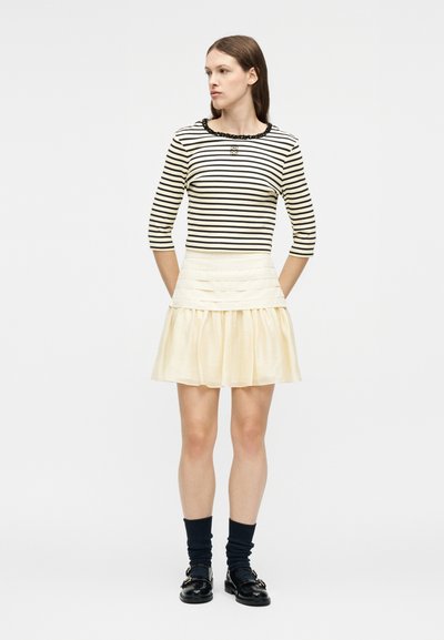 Γυναίκα που στέκεται με τα χέρια πίσω από την πλάτη, φοράει μαύρο-άσπρο ριγωτό τοπ, κρεμ πλισέ φούστα, μαύρες κάλτσες και μαύρα μοκασίνια.