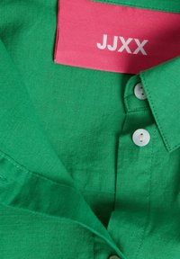 Žalias lininis marškinis su apykakle, su baltomis sagomis ir rausvu etiketu, ant kurio užrašyta "JJXX". Lygi tekstūra, paprasti dizaino akcentai.