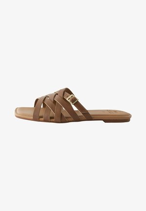 Sandales à semelle plate marron avec des brides croisées, dont une ornée d'une boucle dorée. La semelle est plate et en matériau lisse.