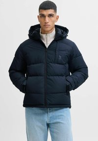 Chaqueta de plumas navy con capucha, con cierre de cremallera en la parte delantera, puños acanalados y secciones cosidas para aislamiento. Combinada con jeans azul claro.