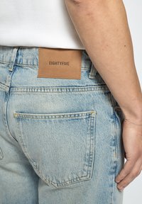 Light blue denim jeans with a faded texture, featuring a brown leather patch labeled "EIGHTYFIVE" on the back. Two rear pockets visible.