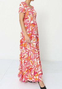 Robe maxi à motifs vifs présentant des tourbillons blancs, roses et oranges, des manches courtes à volants et une jupe à volants. Des chaussures noires complètent le look.