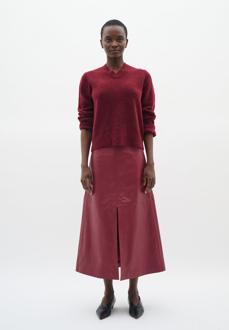 Roter strukturierter Strickpullover mit V-Ausschnitt, kombiniert mit einem knöchellangen roten Lederrock mit einem Schlitz vorne. Schwarze spitze Ballerinas runden den Look ab.