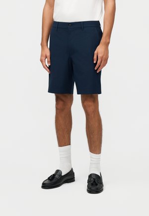 Άνδρας που φοράει ναυτικά μπλε ραμμένα σορτς, λευκές κάλτσες, μαύρα loafers με φούντες και λευκό πουκάμισο φορεμένο μέσα στο παντελόνι, στέκεται μπροστά σε μια απλή υποστήριξη.