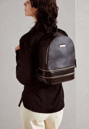Femme portant une chemise noire et un pantalon couleur crème, portant un sac à dos en cuir noir avec des fermetures éclair dorées et une partie inférieure texturée.