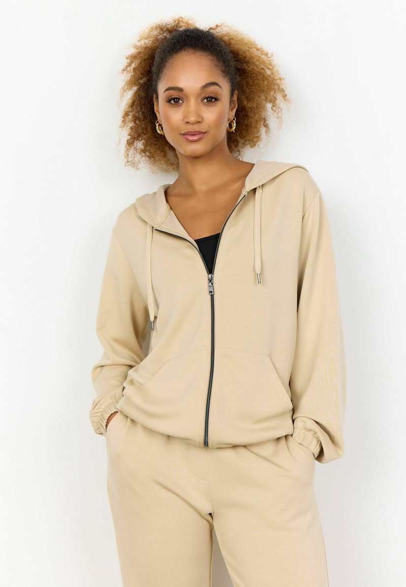 Felpa con zip beige, dotata di cappuccio e due tasche frontali, realizzata in tessuto morbido. Presenta polsini elasticizzati e un design dal taglio comodo.