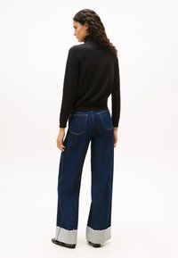 Donna con lunghi capelli ricci che indossa un maglione nero a collo alto e jeans ampi di un blu scuro con orli risvoltati, abbinati a scarpe nere con borchie.