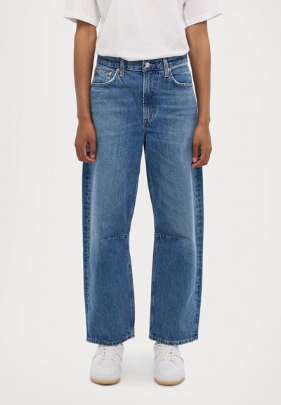 Weite Denim-Jeans in Mittelblau mit hoher Taille, subtiler Ausbleichung und klassischem Fünf-Taschen-Design. Kombiniert mit weißen Sneakers.