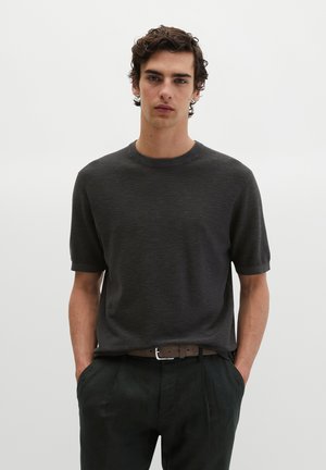 Jeune homme aux cheveux courts et bouclés portant un pull à manches courtes gris foncé et un pantalon noir avec une ceinture marron, les mains dans les poches, fond neutre.