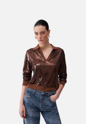 Donna con capelli pettinati all'indietro indossa una maglia marrone a maniche lunghe con paillettes, dettagli su collo e polsini, abbinata a jeans blu con motivo, in piedi con una mano in tasca.