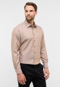 Hellbraune Langarm-Hemdbluse mit einer Tasche, aus einem glatten Stoff, mit Kragen und dunklen Knöpfen. Getragen mit schwarzen Hosen.
