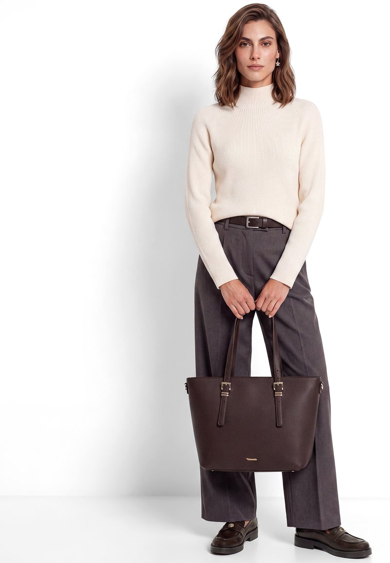 Femme debout tenant un sac cabas en cuir marron foncé, portant un pull côtelé crème, un pantalon gris à jambes larges et des mocassins foncés sur fond blanc.