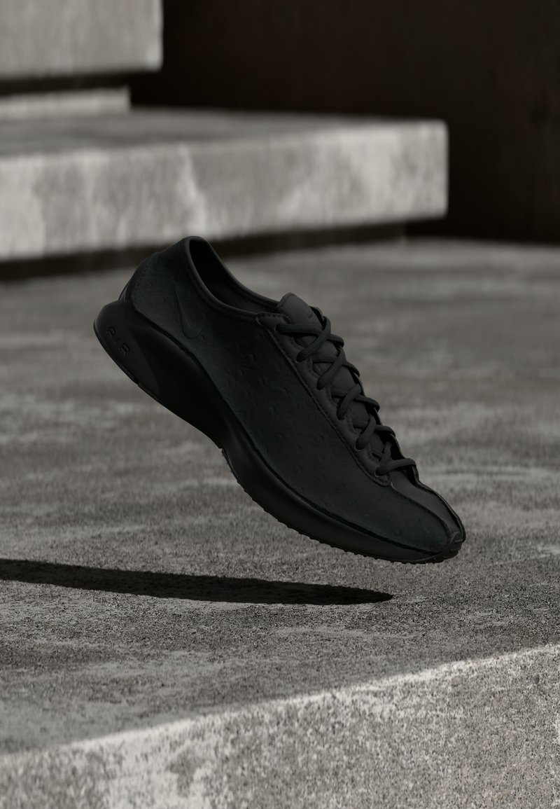Μαύρο αθλητικό παπούτσι με λεία υφή, στρογγυλεμένη μύτη και επίπεδη σόλα. Διαθέτει διακριτικό λογότυπο και παραδοσιακό σύστημα κορδονιών, αιωρούμενο πάνω από σκυρόδεμα.