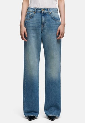 Person iført løstsiddende, højtsiddende blå jeans med falmede områder, kombineret med sorte spidse sko og en hvid top.