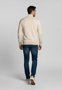 Suéter beige de punto con cuello alto, combinado con jeans azul oscuro y zapatillas claras. El ajuste es entallado y la textura parece suave.