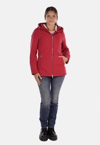 Giacca impermeabile rossa con cappuccio, chiusura con zip, tasche laterali e rivestimento interno morbido. Abbinata a jeans blu scuro e scarpe nere.