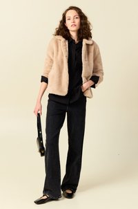 Beige nepbont jas met een kraag, gedragen over een zwart shirt. Gecombineerd met zwarte flare jeans en zwarte ballerina's, met een schoudertas.