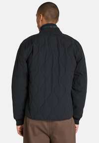 Svart vadderad jacka med en ribbad krage och ett rutmönster i grönt och blått. Designen har detaljer med böjda sömmar.