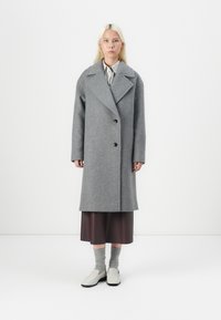 Cappotto di lana grigio con ampio revers, design doppiopetto e bottoni neri. Indossato sopra una gonna marrone con calzini grigi e scarpe chiare.