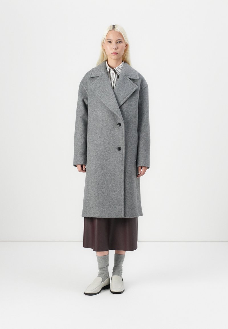 Cappotto di lana grigio con ampio revers, design doppiopetto e bottoni neri. Indossato sopra una gonna marrone con calzini grigi e scarpe chiare.