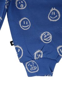 Blauwe gebreide trui met verschillende bedrukte glimlachende en fronsende gezichten in het wit, met geribbelde manchetten. Zwarte label aan de zijkant.