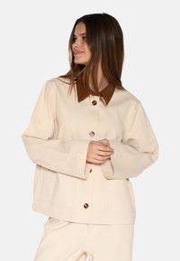 Crème-kleurige jas van gestructureerde stof, met een leren kraag in bruin en oversized knopen. Wijde mouwen en een relaxed fit.