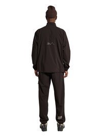 Person iført sort jakke med hvid omrids grafik på ryggen, sorte bukser med teksten "223" på venstre ben, brun hue og grå sneakers, set bagfra.