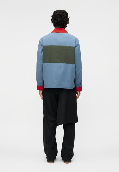 Άτομο με σγουρά μαλλιά που φορά μπλε μπουφάν με πράσινη επιφάνεια, κόκκινο γιακά και μανσέτες, μαύρο παντελόνι με στρώσεις υφάσματος στο πίσω μέρος.