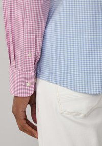 Koszula w kratę typu gingham z różowymi i niebieskimi sekcjami, z guzikami przy mankietach oraz miękkim, lekkim materiałem. Częściowo widoczne białe spodnie.