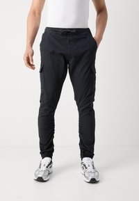 Zwarte cargobroek met verstelbare taille, taps toelopende enkels, zijzakken en een lichtgewicht stof, gecombineerd met zilveren sportschoenen.