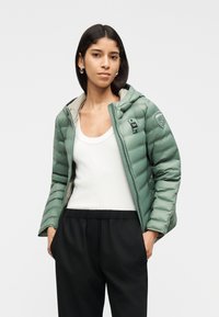 Γυναίκα με ανοιχτό πράσινο μπουφάν τύπου puffer πάνω από λευκή μπλούζα χωρίς μανίκια και μαύρο παντελόνι, με τα χέρια στις τσέπες, ποζάροντας σε απλό φόντο.