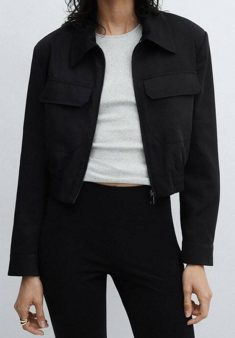 Femme portant une veste noire courte avec poches à rabat sur un crop top gris clair côtelé et un pantalon noir taille haute.
