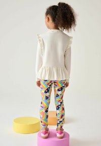 Bílé tričko s dlouhými rukávy, rýhovanými rukávy a peplum lemem, spárované s barevně duhovými legínami a růžovými botami.