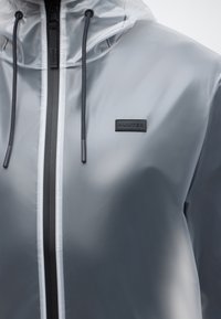 Διαφανές γκρι αδιάβροχο μπουφάν με μαύρο φερμουάρ, μαύρα κορδόνια και μαύρη πλακέτα με το λογότυπο "HUNTER" στο αριστερό στήθος.